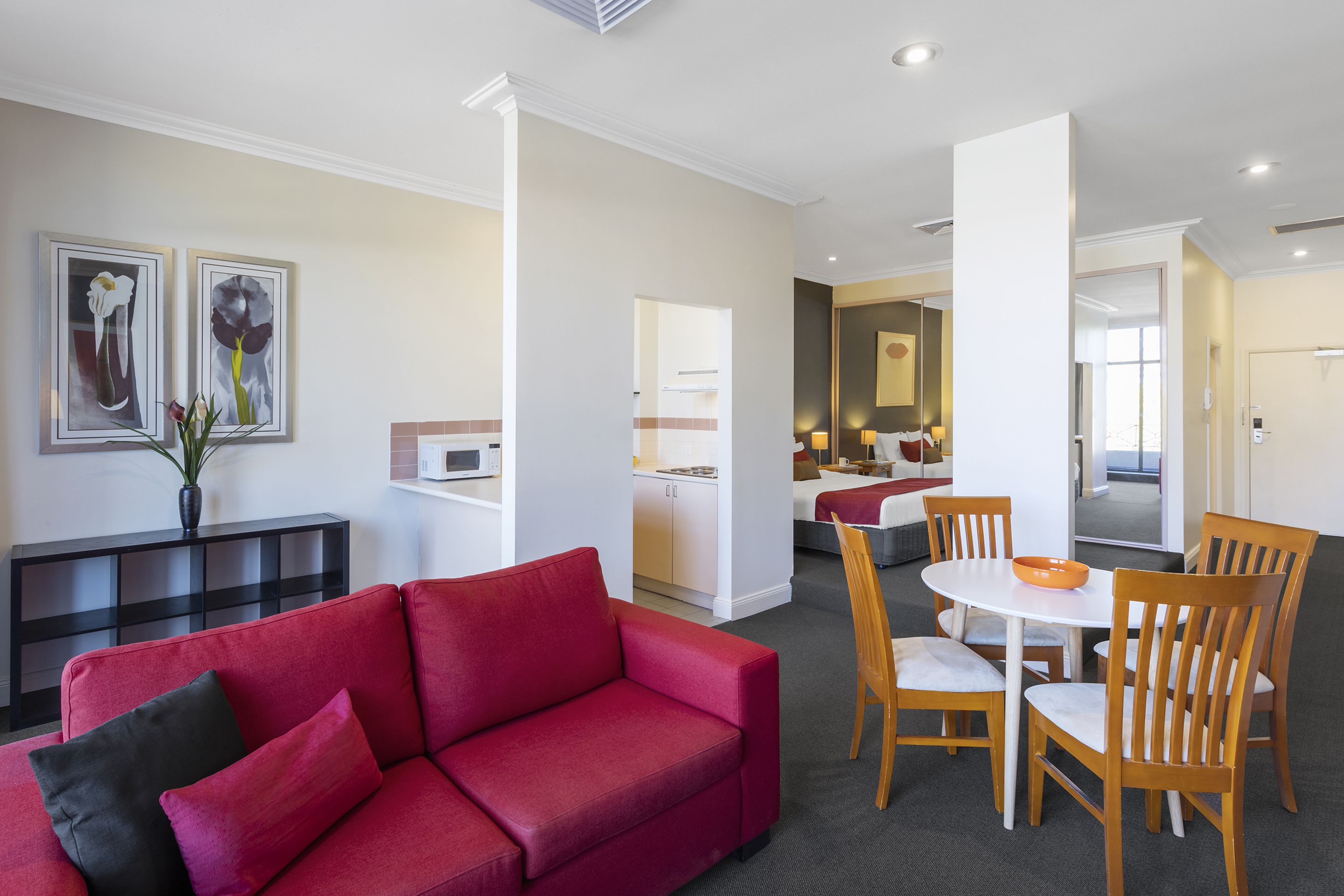 A modern living room with a red sofa, wooden dining chairs, and a small table. There is a kitchen area in the background with white cabinets and a dining table.