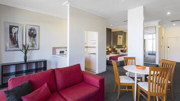 Una sala moderna con un sofá rojo, sillas de comedor de madera y una pequeña mesa. En el fondo, se distingue una zona de cocina con armarios blancos y una mesa de comedor.