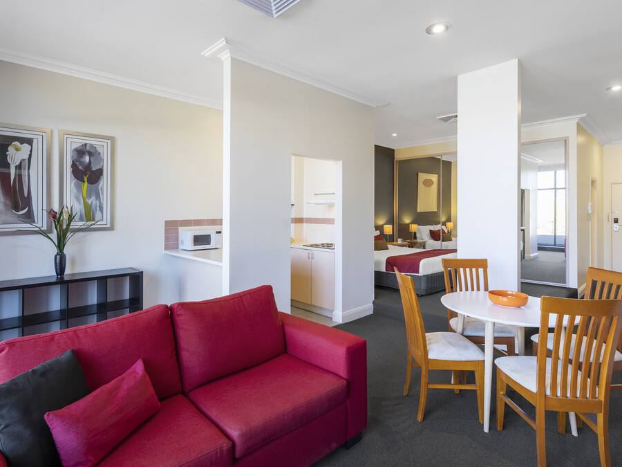 Una sala moderna con un sofá rojo, sillas de comedor de madera y una pequeña mesa. En el fondo, se distingue una zona de cocina con armarios blancos y una mesa de comedor.