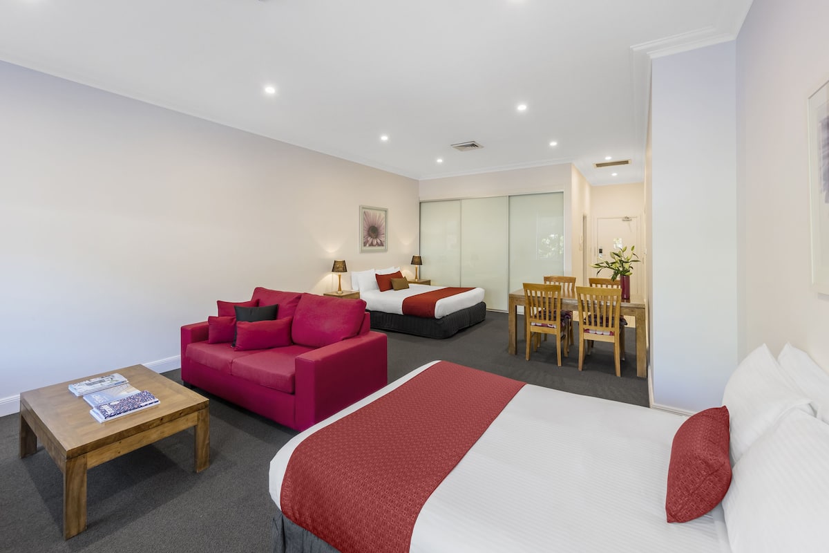 Una habitación de hotel con una cama, un sofá rojo, una mesa de centro de madera y un escritorio con sillas.