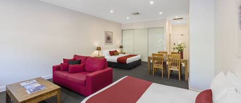 A hotel room with a bed, a red sofa, a wooden coffee table, and a desk with chairs.
