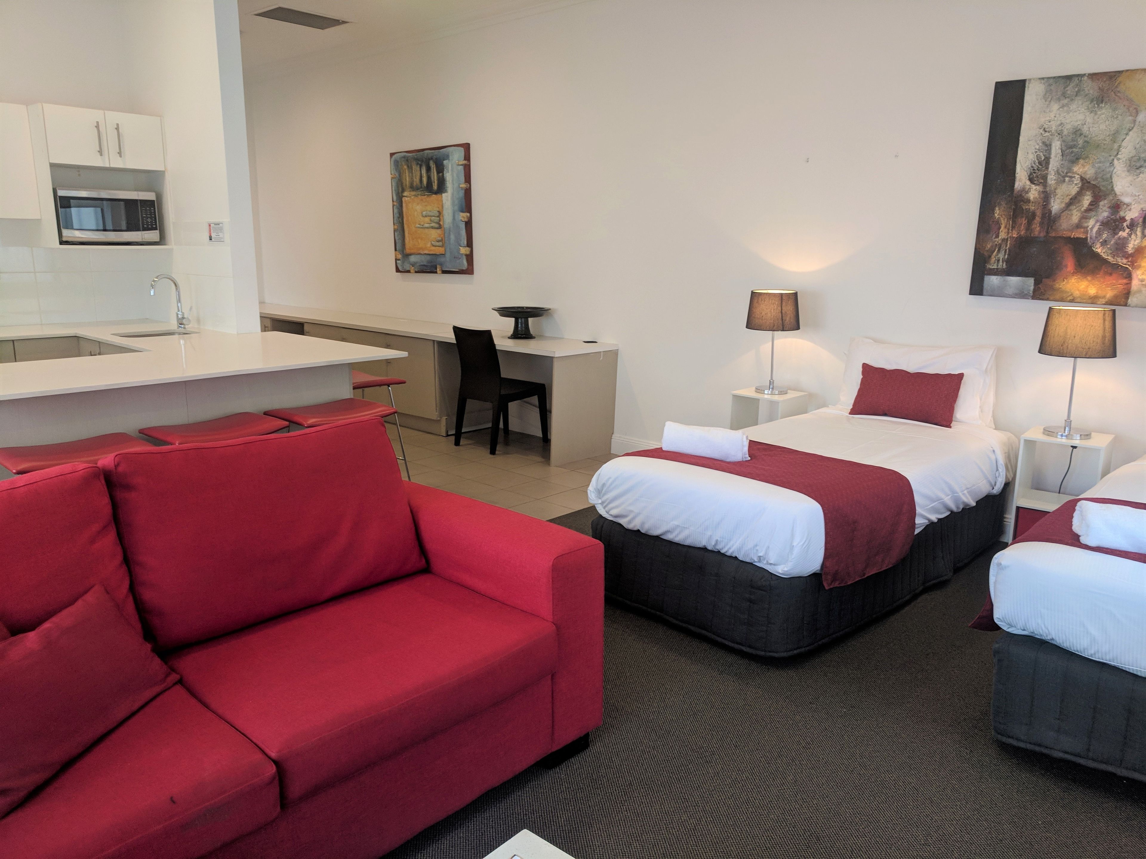 A hotel room with a red sofa, two beds with white linens, a desk, a chair, a microwave, a painting, and a lamp.