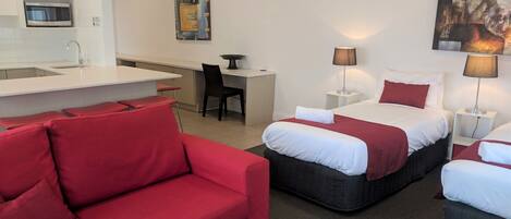 Habitación de hotel con sofá rojo, dos camas con ropa blanca, un escritorio, una silla, microondas, un cuadro y una lámpara.