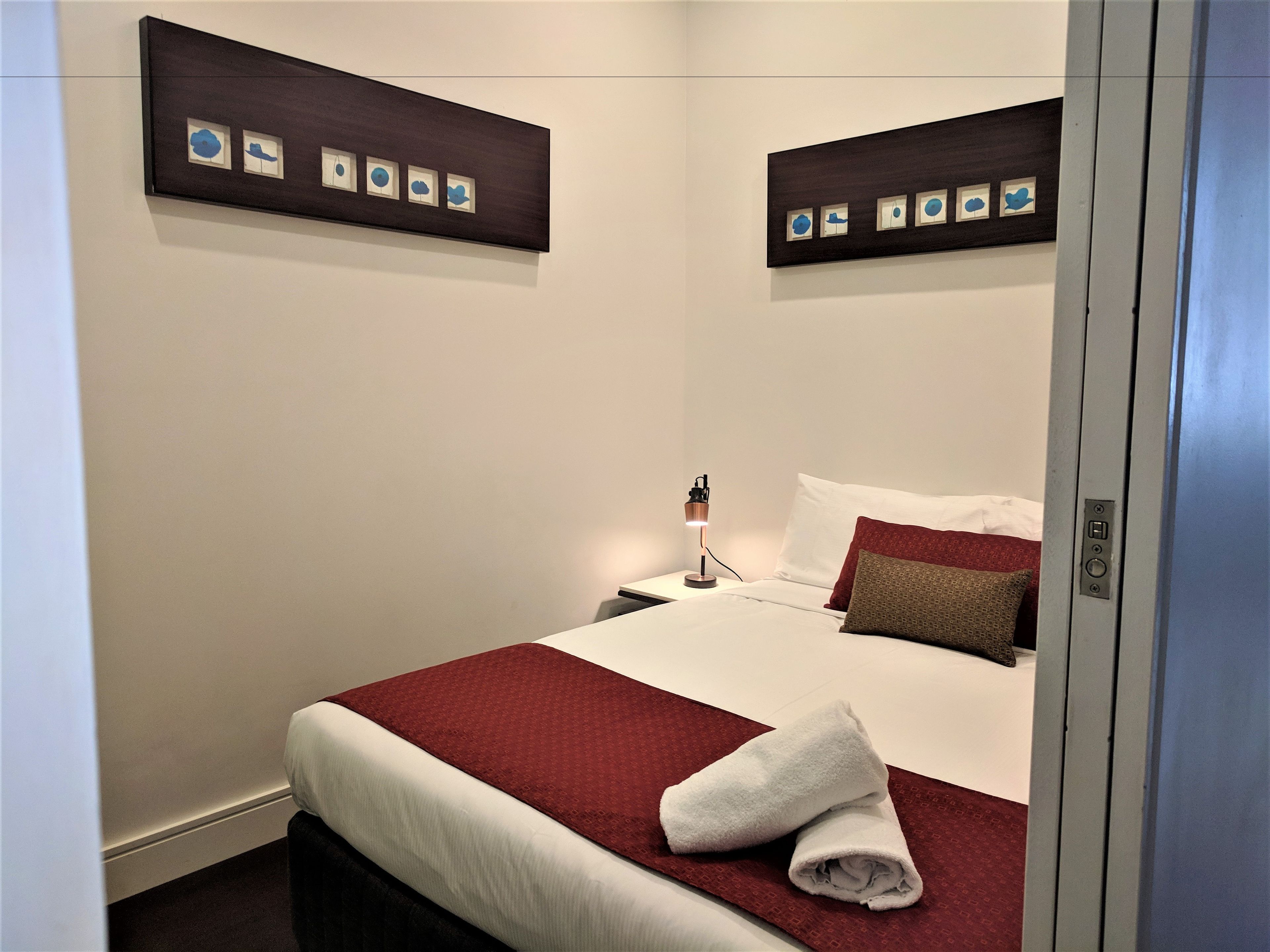 A hotel room with a neatly made bed, a red throw blanket, a white towel, and a nightstand with a lamp.