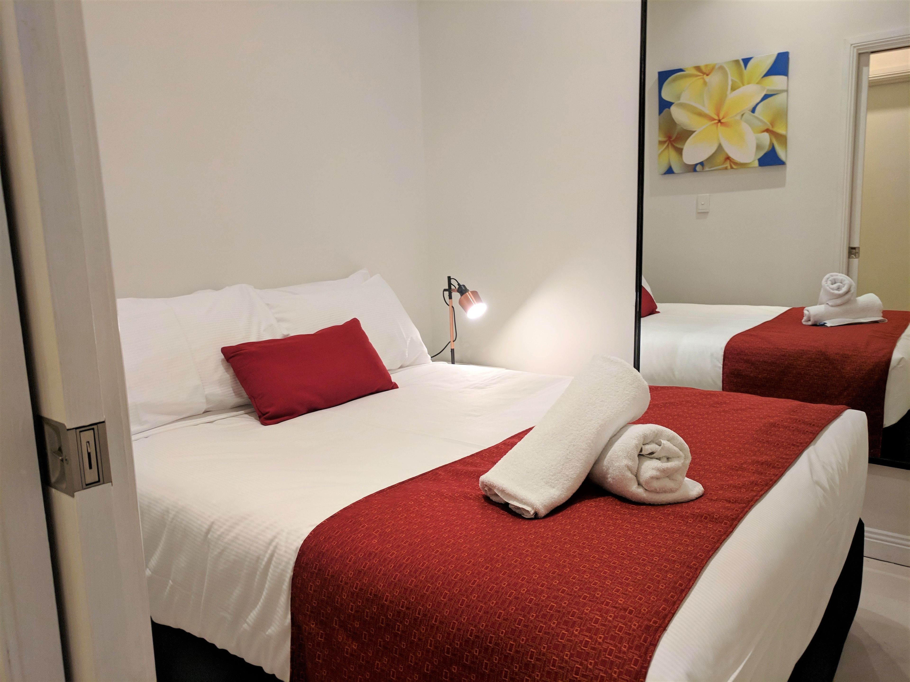 A hotel room with two beds, each with white bedding and red accents. There is a red pillow on one bed, a white towel on the other, and a painting of flowers on the wall.