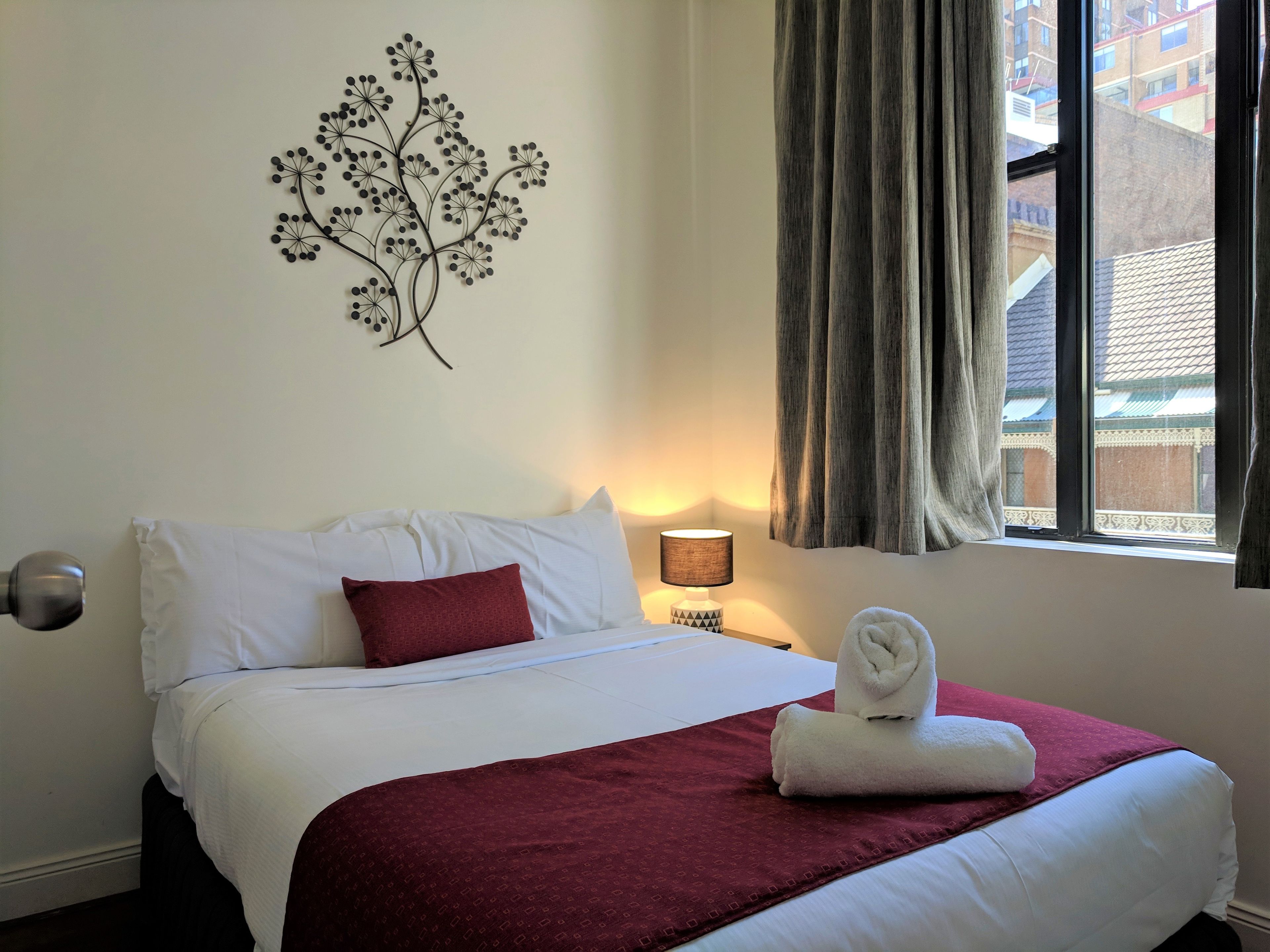 A hotel room with a bed, a decorative wall piece, a nightstand with a lamp, and a window with curtains.