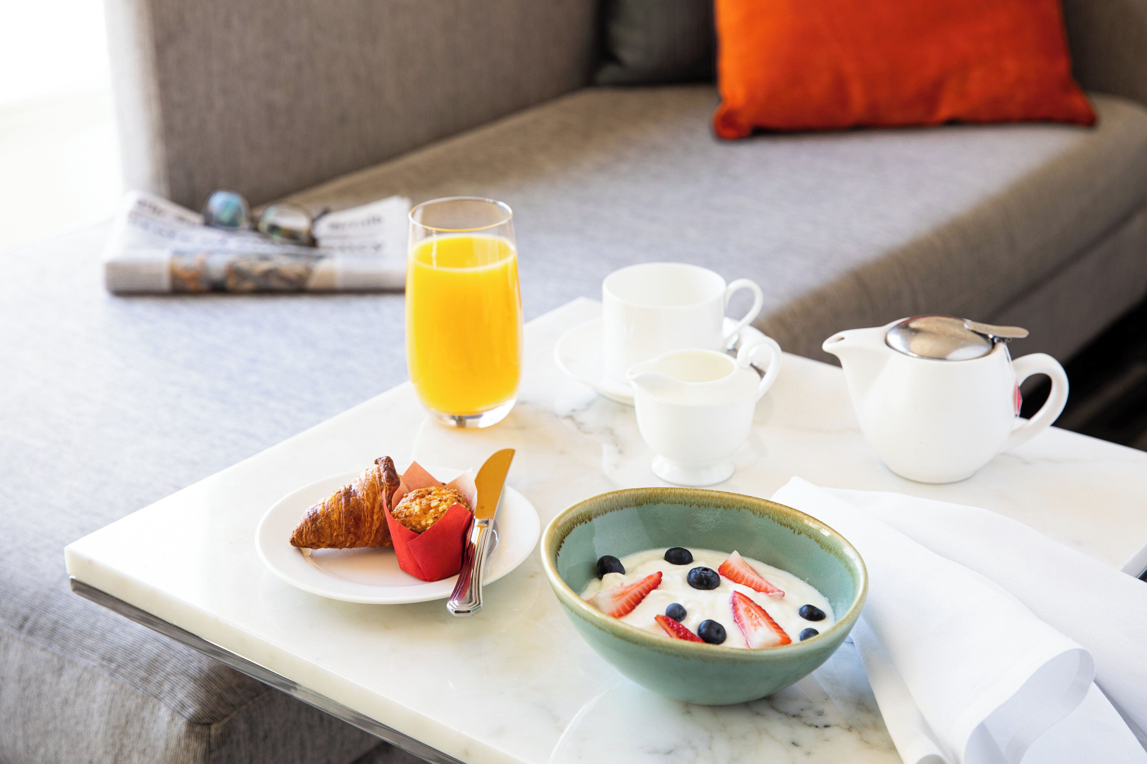 A breakfast setup on a table with croissants, a bowl of yogurt with berries, a glass of orange juice, and a coffee pot.