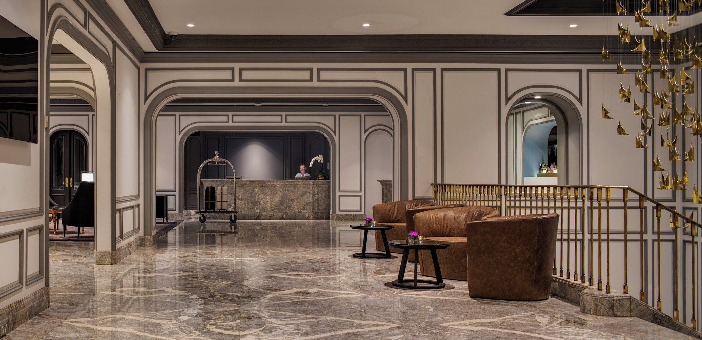A hotel lobby with marble flooring, a reception desk, leather seating, and a staircase with golden railings.