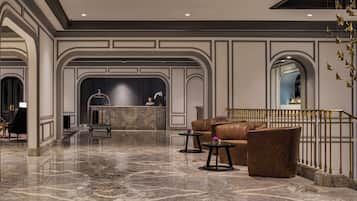 A hotel lobby with marble flooring, a reception desk, leather seating, and a staircase with golden railings.