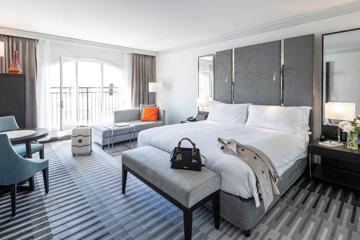 A modern hotel room with a large bed, a desk, a chair, a sofa, and a balcony view.