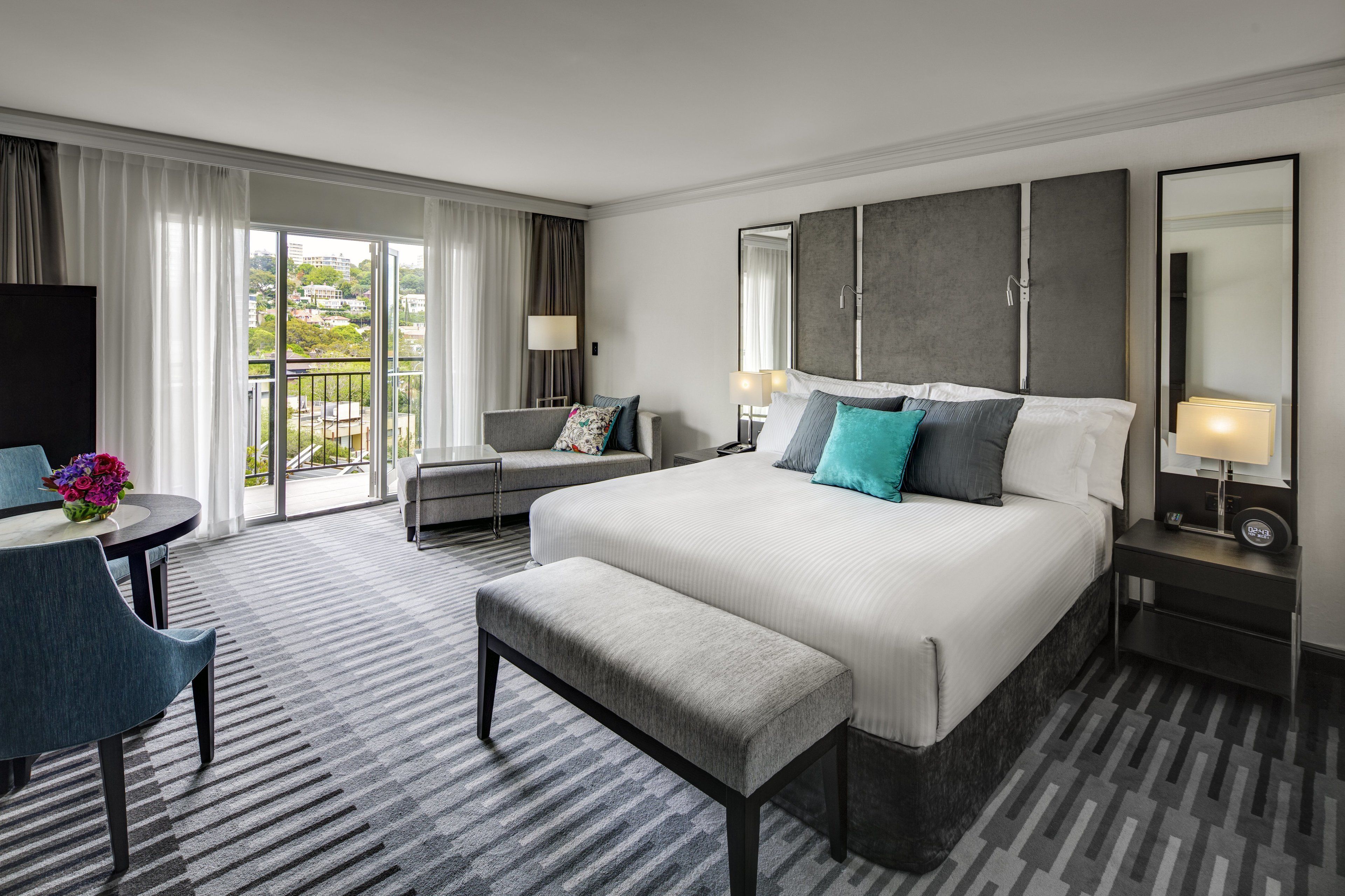 A hotel room with a large bed, a desk, a chair, a small table with a vase of flowers, and a balcony with curtains.