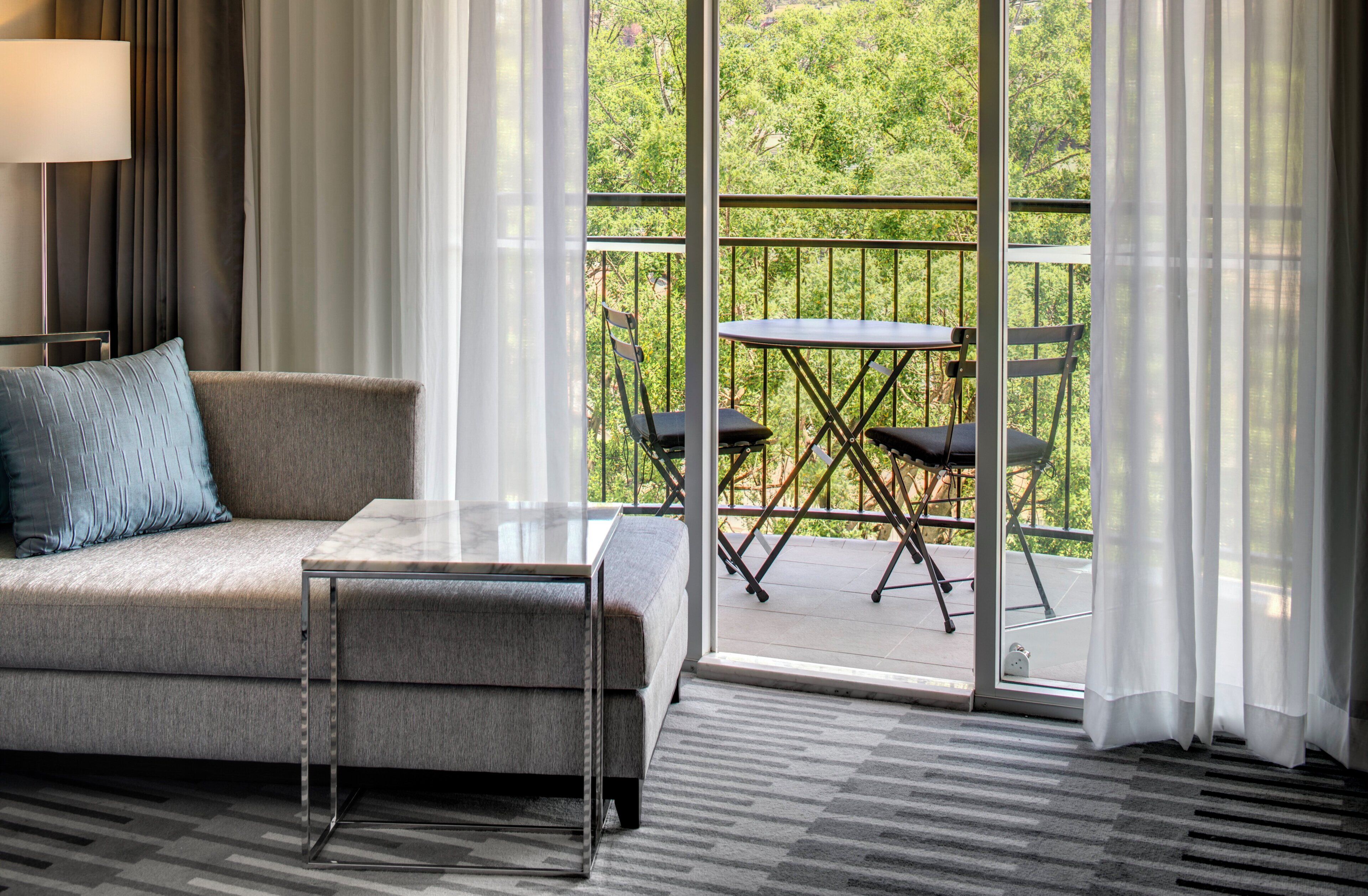 Habitación de hotel moderna con sofá, una mesita, un balcón con mesa y sillas, y vistas a zonas verdes.