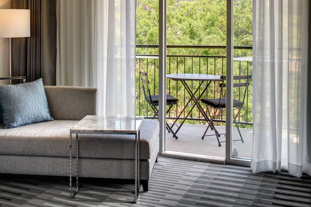 A modern hotel room with a sofa, a small table, a balcony with a table and chairs, and a view of greenery.