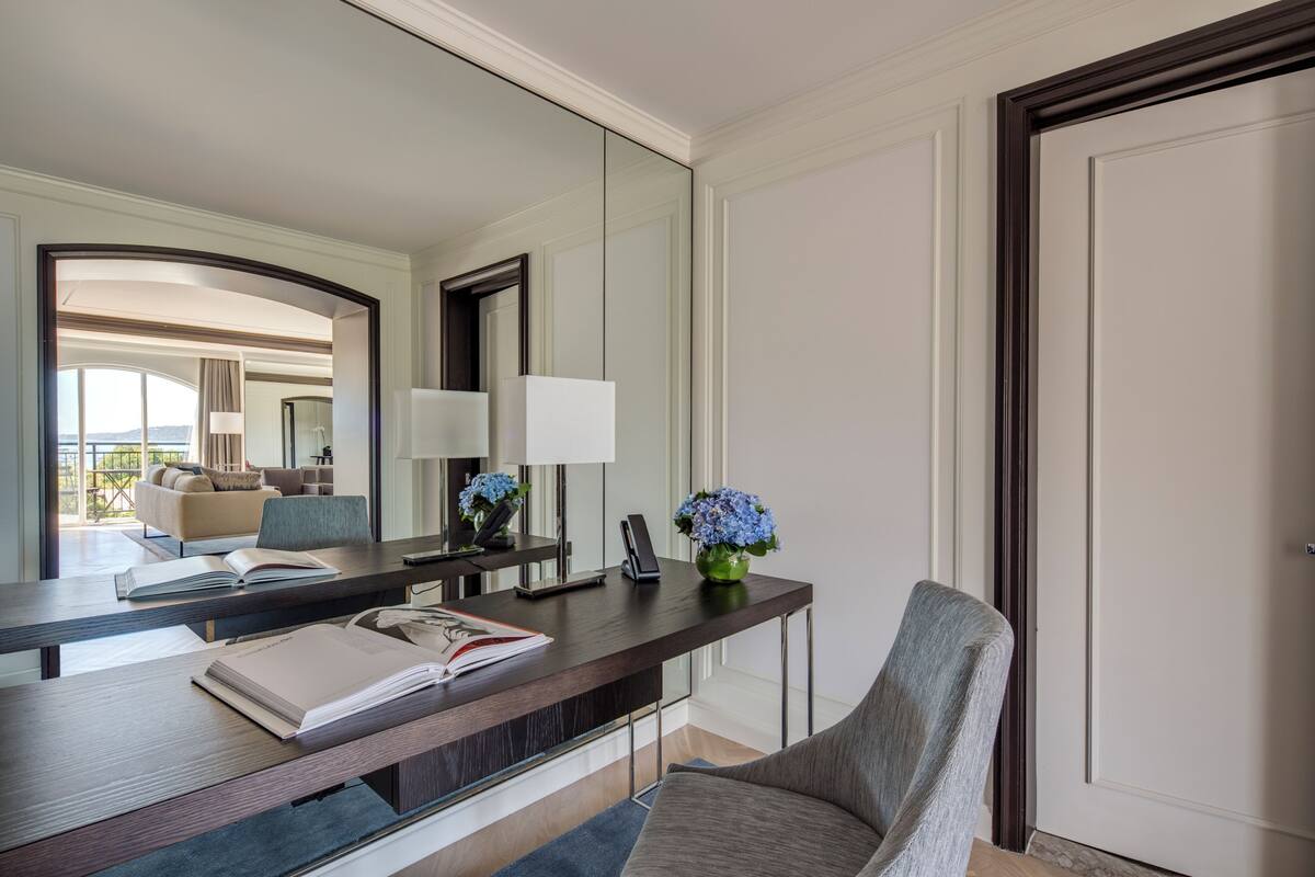 A modern hotel room with a large mirror, a desk with a book, a vase with flowers, and a chair.