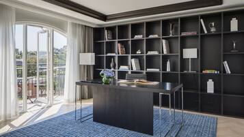 Ein modernes Interieur mit einem zentralen dunklen Holztisch, einem Bücherregal voller Bücher und Dekorationsgegenständen sowie Blick auf eine Stadtlandschaft durch große Fenster.