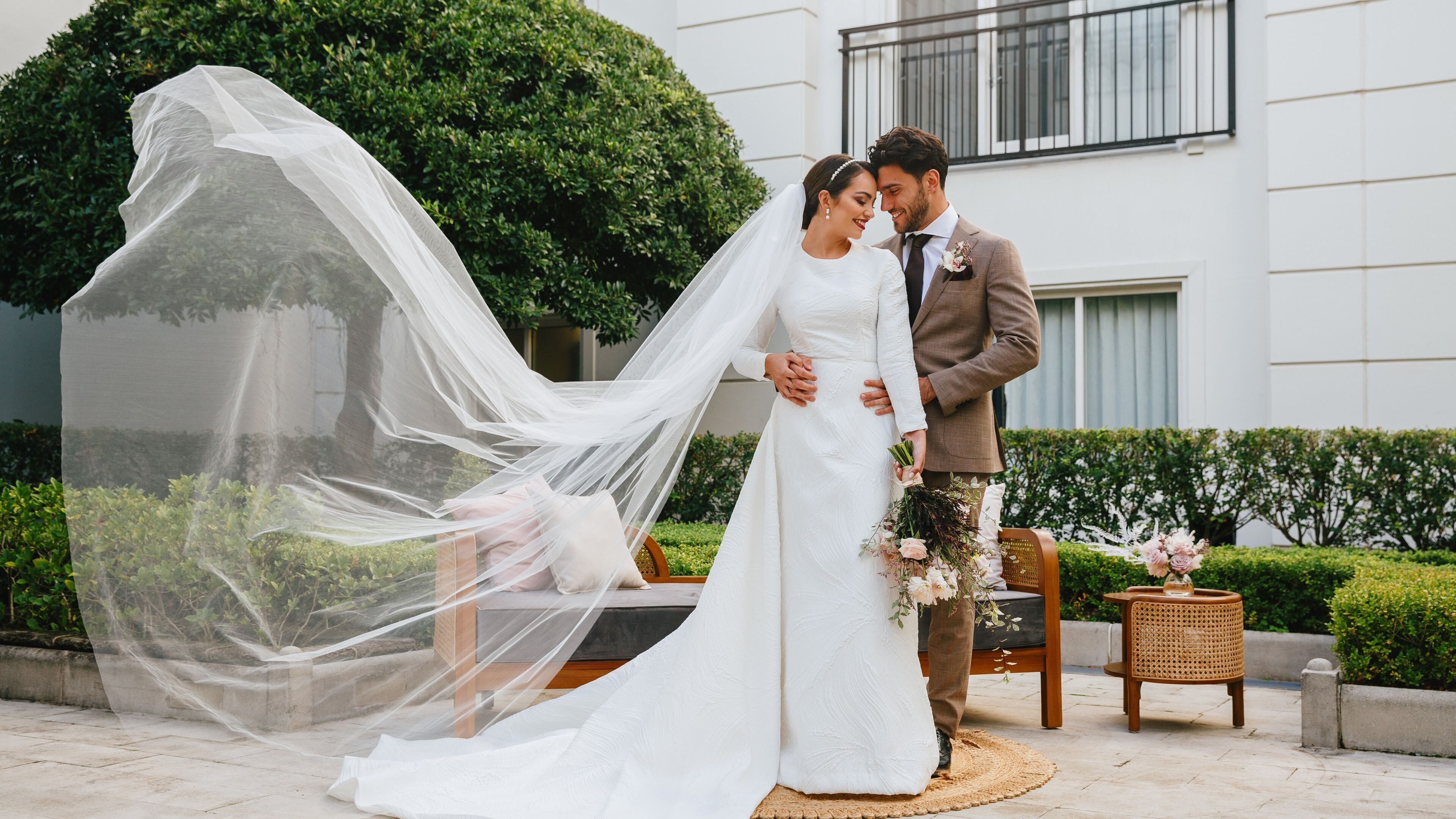 Una pareja vestida de novios al aire libre, con un edificio de fondo.