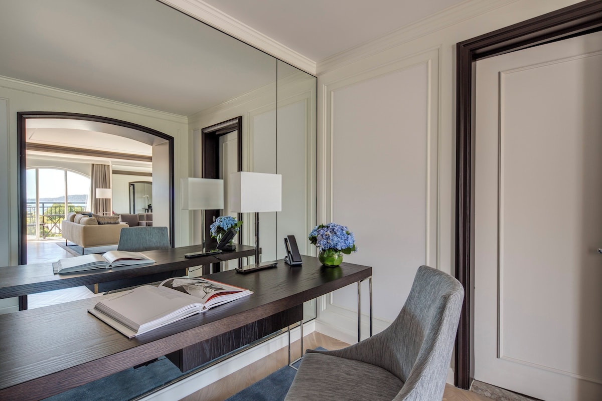 A hotel room with a large mirror, a desk with a book, a vase with flowers, and a chair.