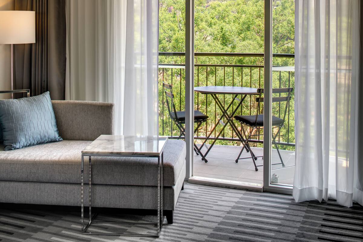 A modern hotel room with a sofa, a small table, and a balcony with chairs.