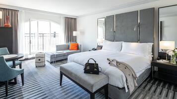A modern hotel room with a large bed, a desk, a chair, a suitcase, and a balcony view.
