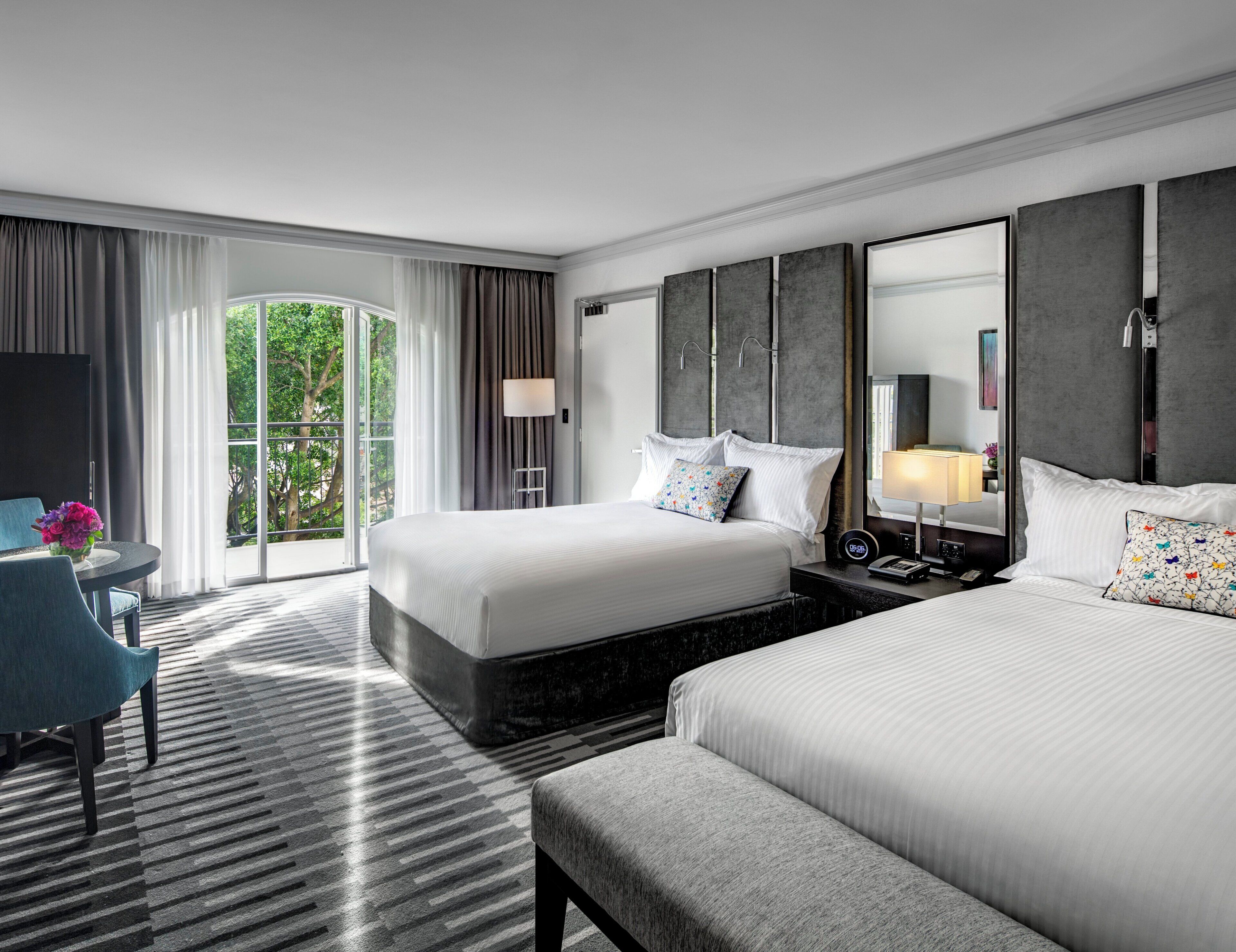 Habitación de hotel con una cama grande, una cama más pequeña, un escritorio con silla, un ventanal con cortinas y vistas al exterior con vegetación.
