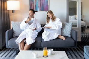 A couple in white robes sitting on a grey sofa in a hotel room with a coffee table, a lamp, and a painting on the wall.