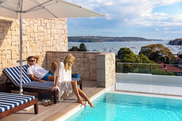Zona junto a la piscina con pared de piedra, sillas de descanso y vista al mar.