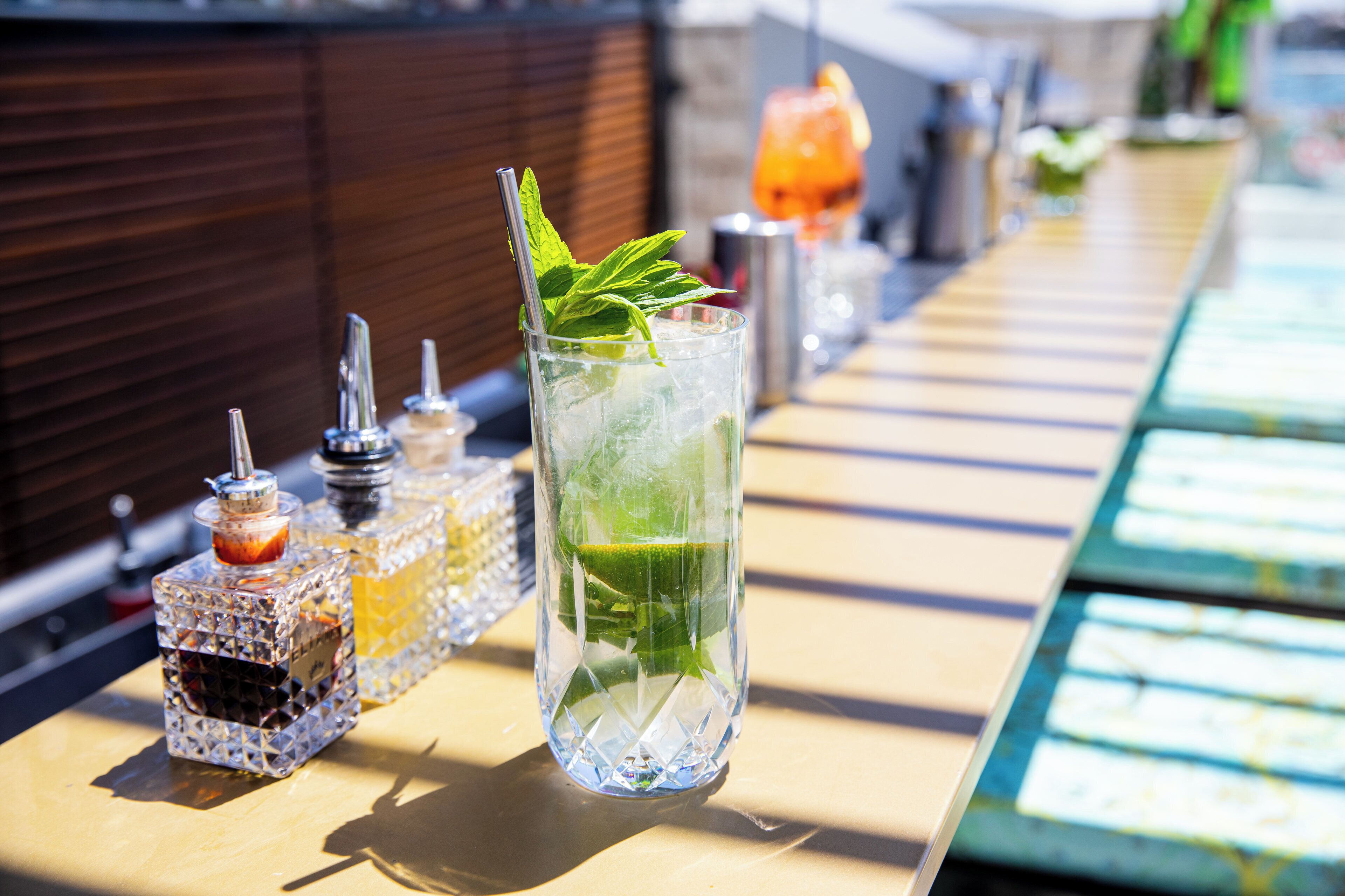 Un coin bar avec un verre contenant une boisson claire, décorée de feuilles de menthe, de glaçons et d’une tranche de citron vert.