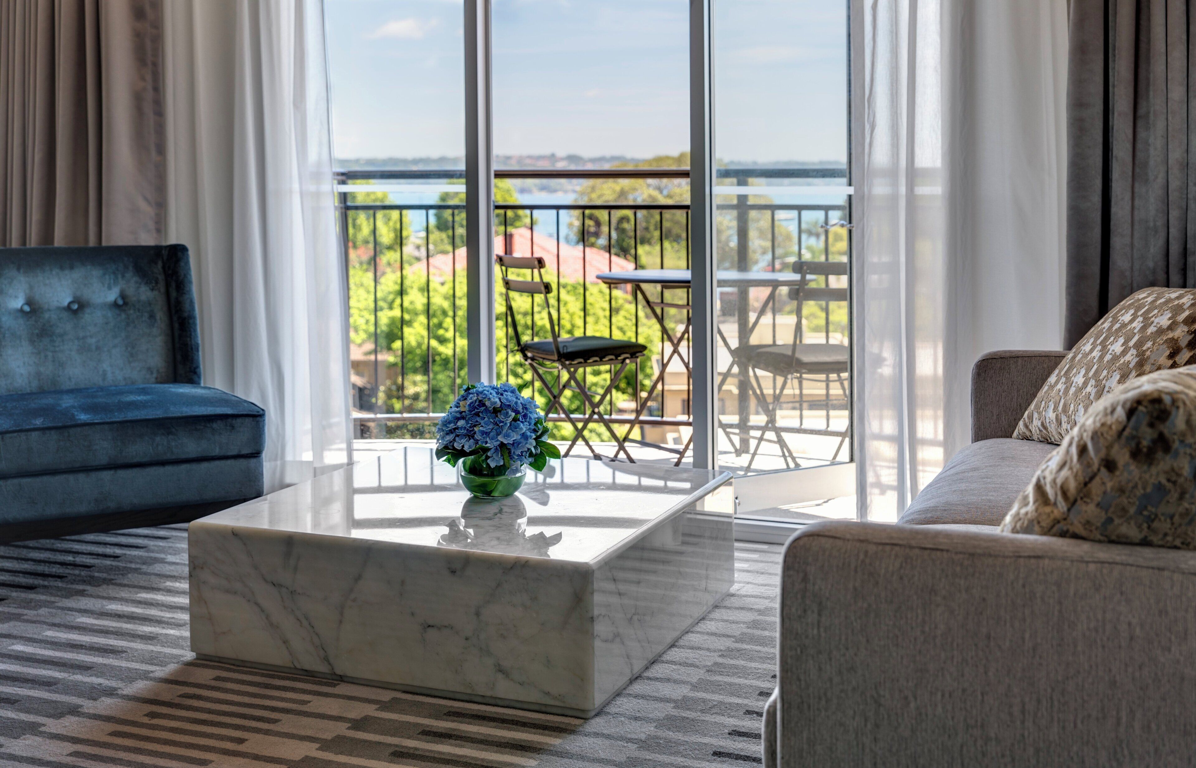 A modern living room with a marble coffee table, a blue sofa, and a grey sofa with patterned cushions. A balcony with a view of greenery and buildings is visible through large windows.