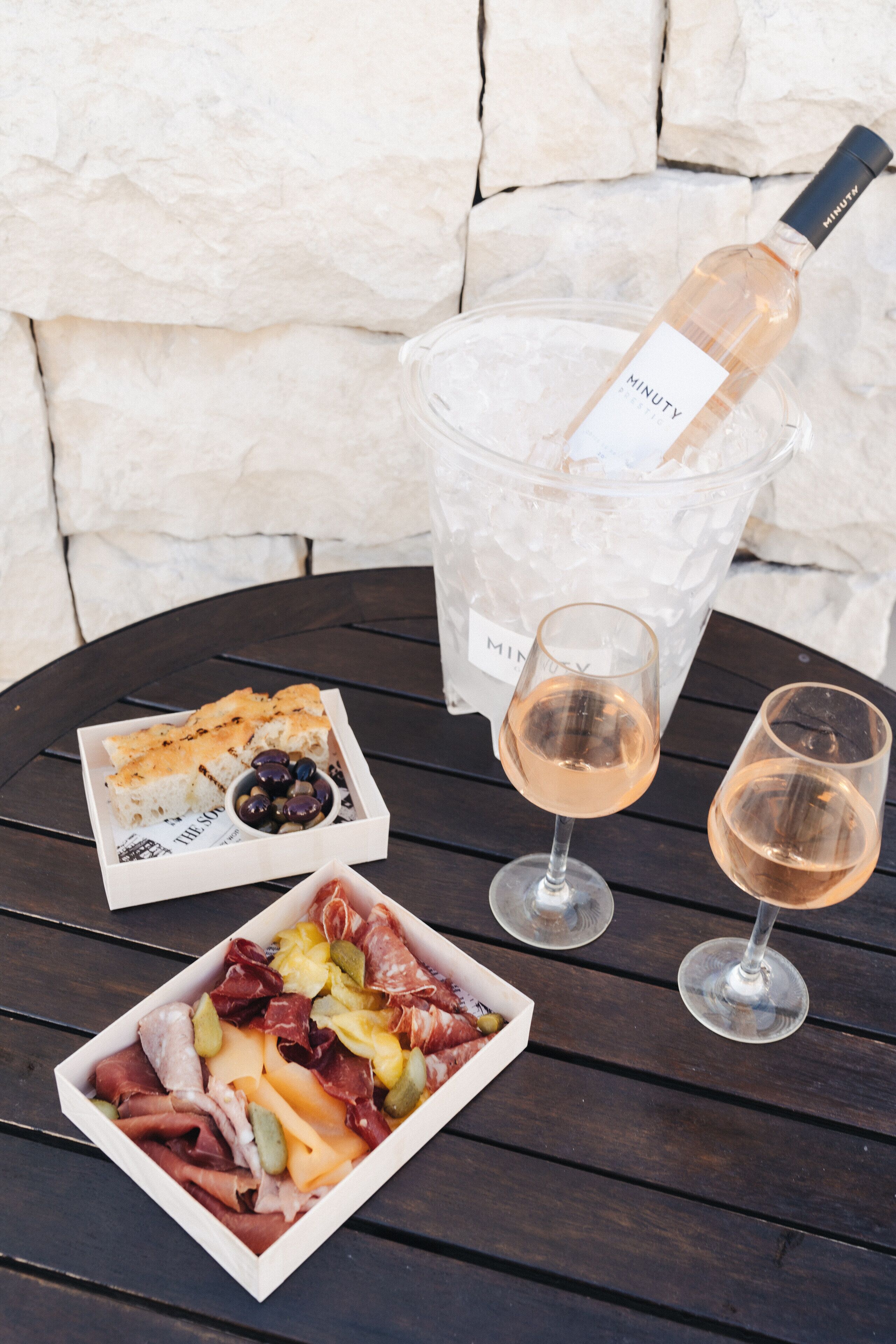 A table with a bottle of wine, two glasses, a box of charcuterie, and a basket of olives.