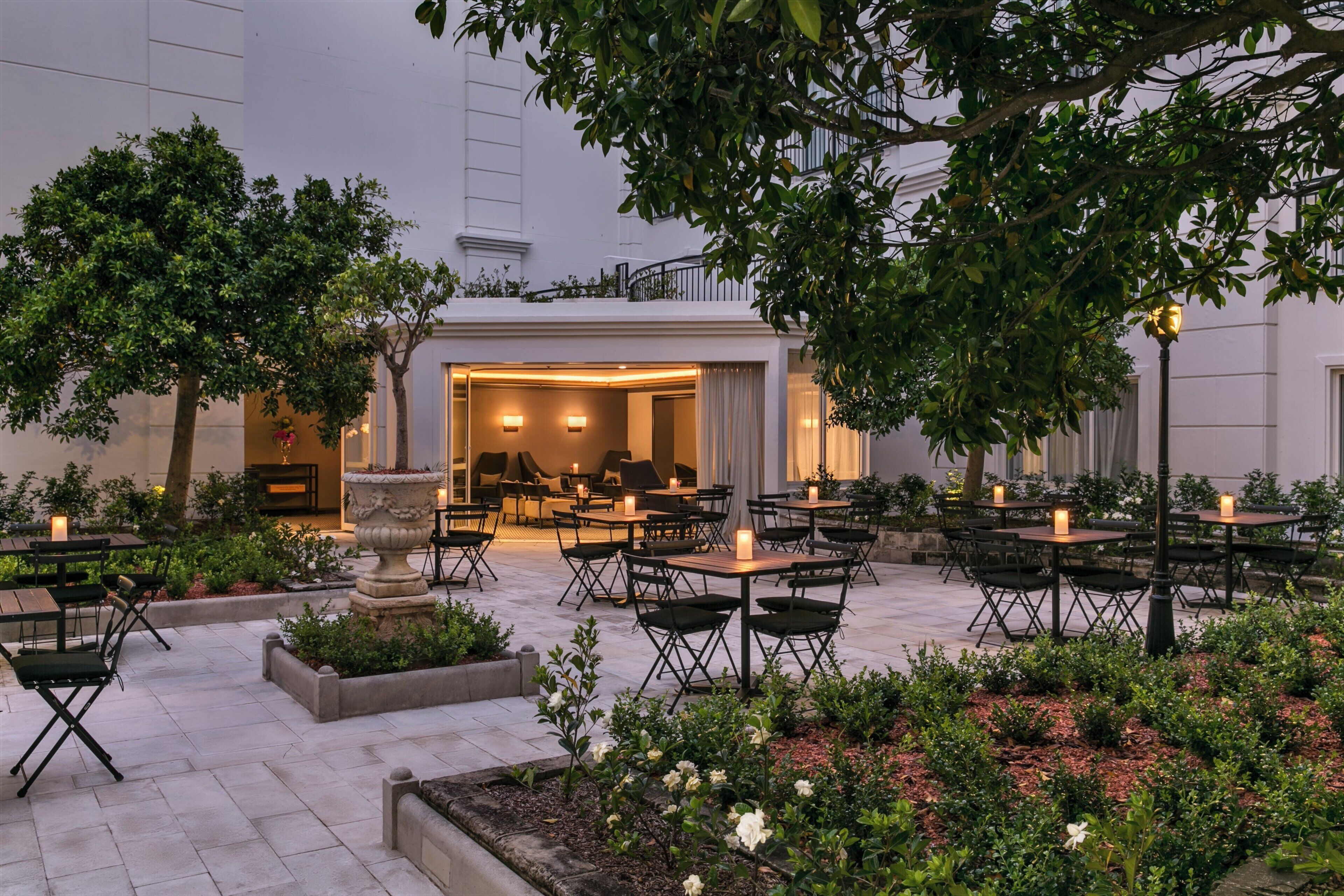 Un espace extérieur aménagé avec des tables et des chaises, entouré de verdure et situé près de l’entrée d’un bâtiment.