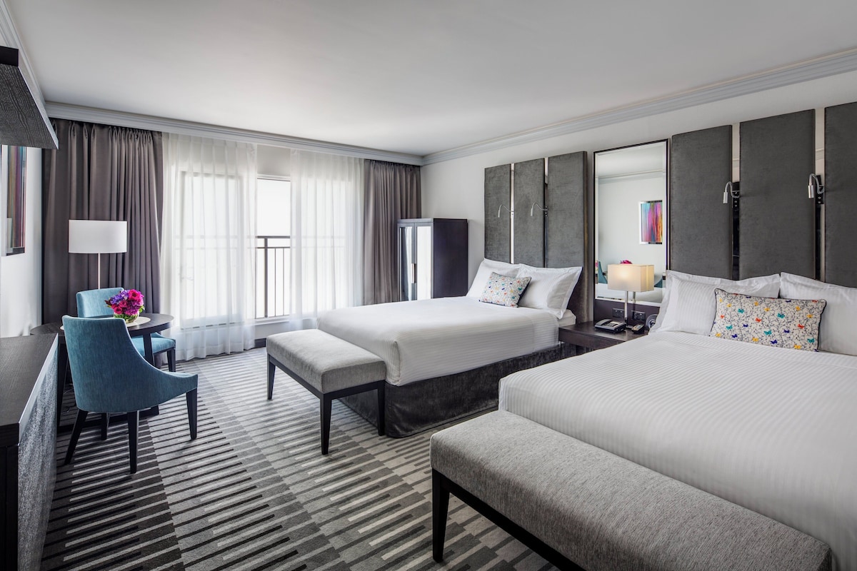 A hotel room with two beds, a desk, a chair, a lamp, and a large window with curtains.