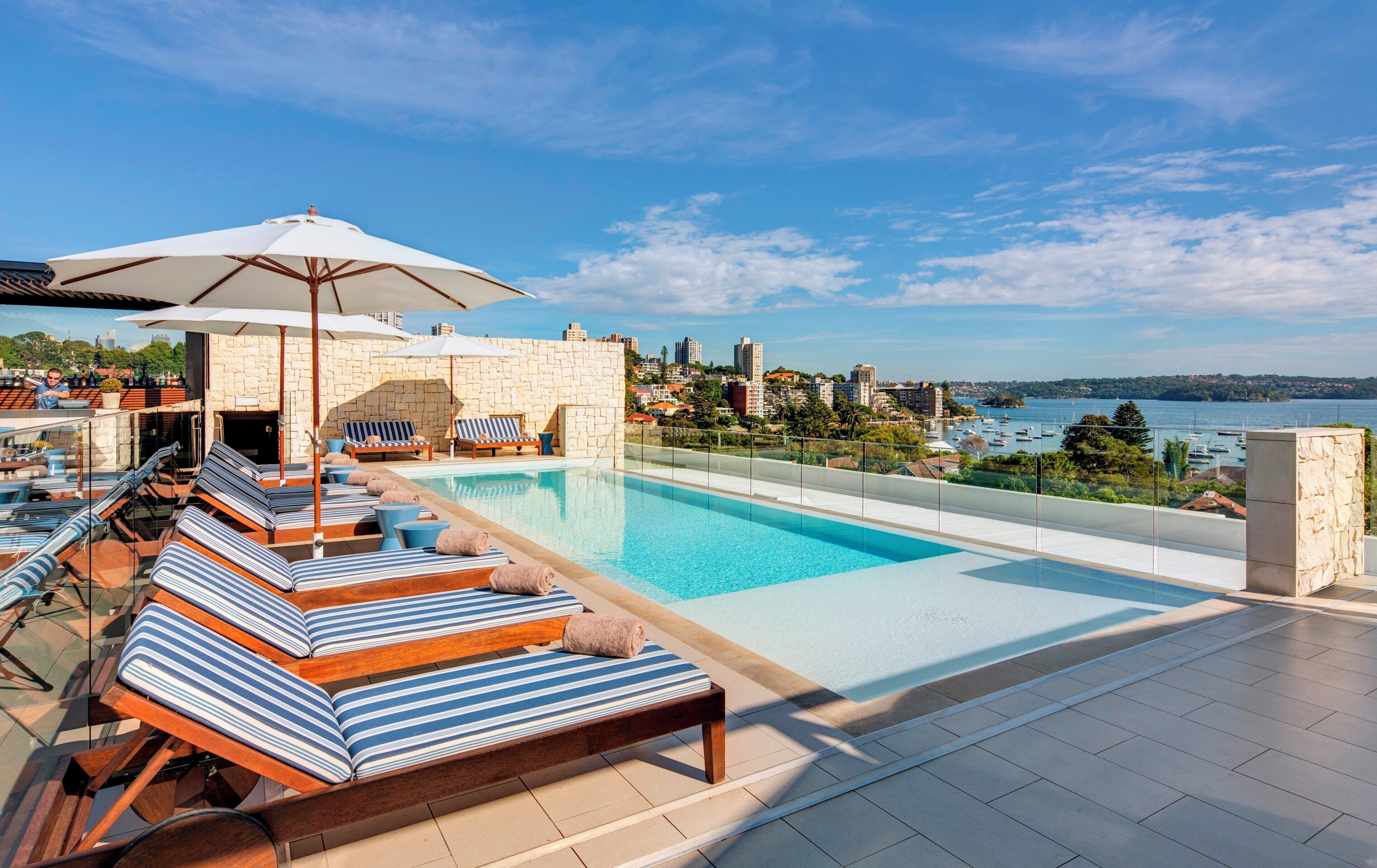 A rooftop pool area with lounge chairs and umbrellas, overlooking a cityscape and body of water.