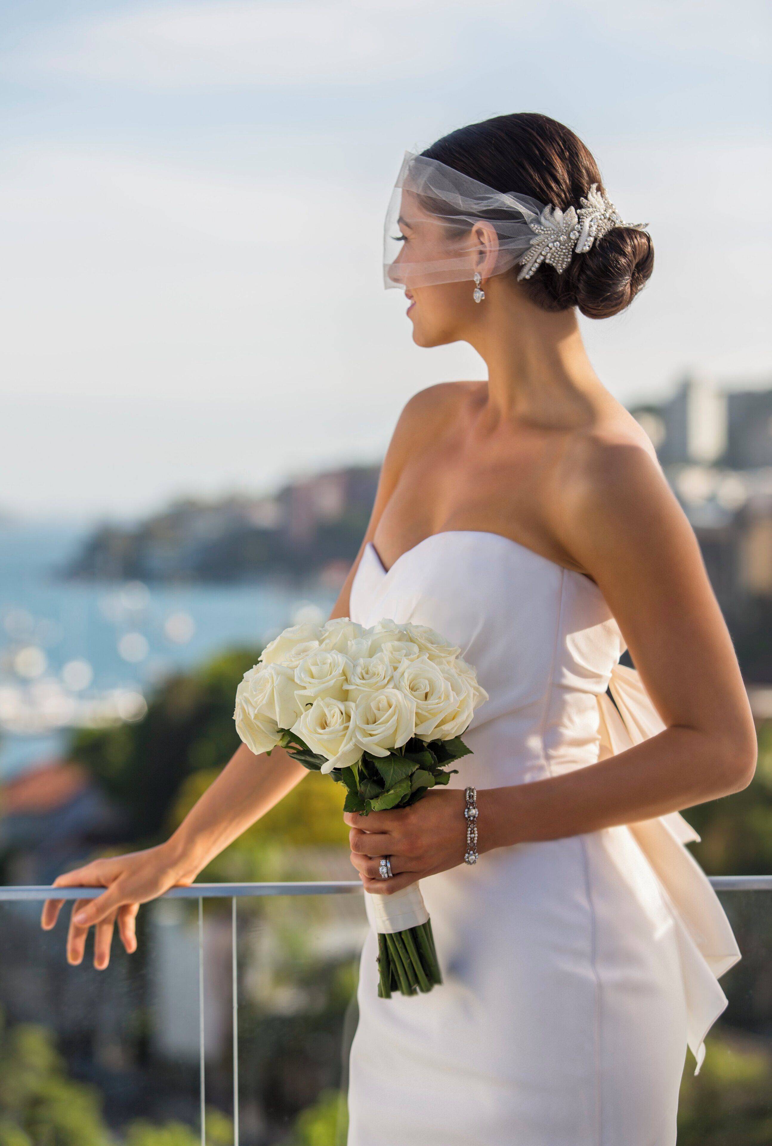 Una persona con un vestido blanco sostiene un ramo de rosas blancas, de pie en un balcón con vista a la ciudad.