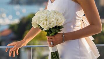 Una persona con un vestido blanco sostiene un ramo de rosas blancas, de pie en un balcón con vista a la ciudad.