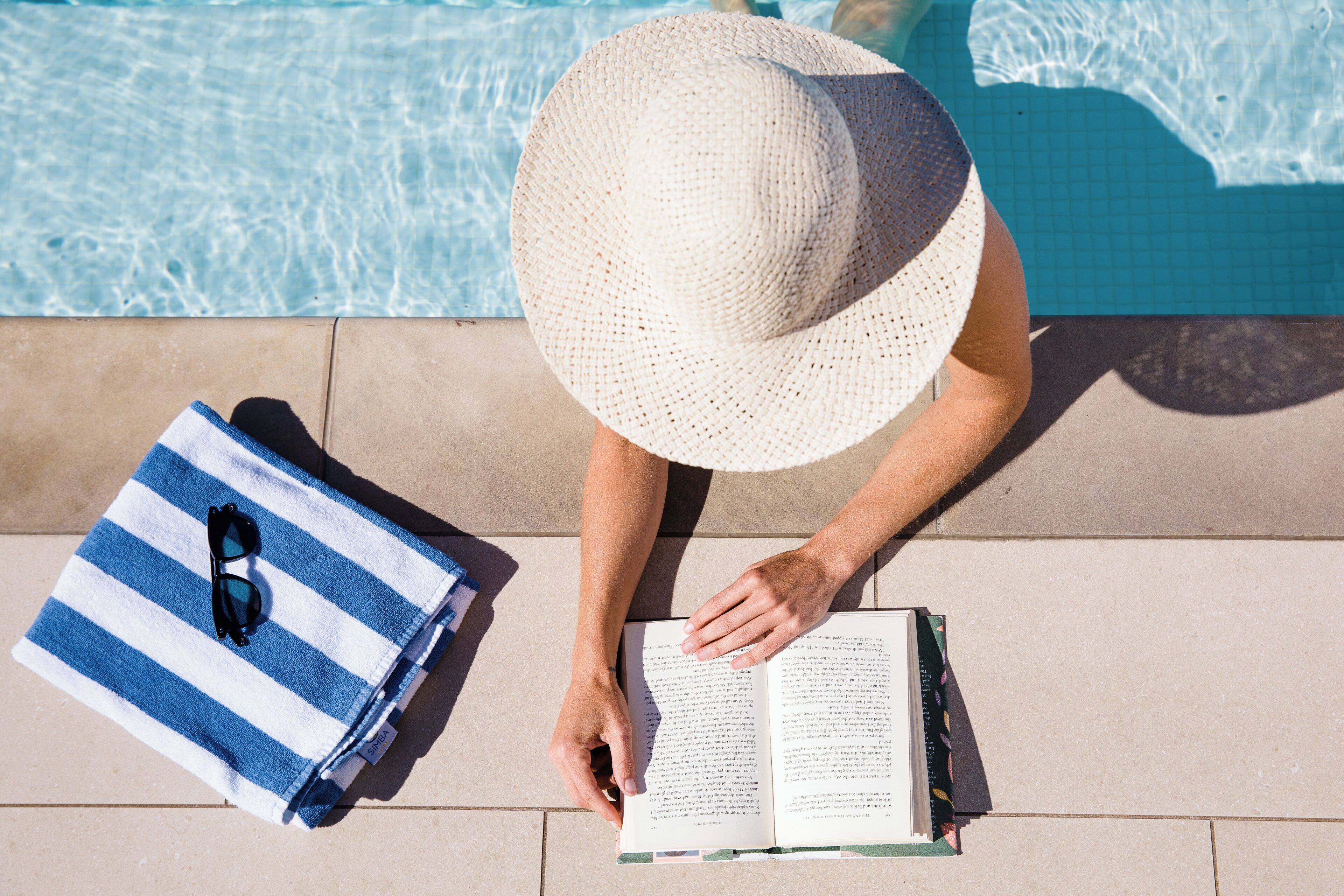 A person reading a book by a poolside with a striped towel and sunglasses on it.