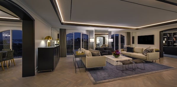 A modern living room with a sofa, coffee table, and chairs. There is a large window with a view of the city at night.