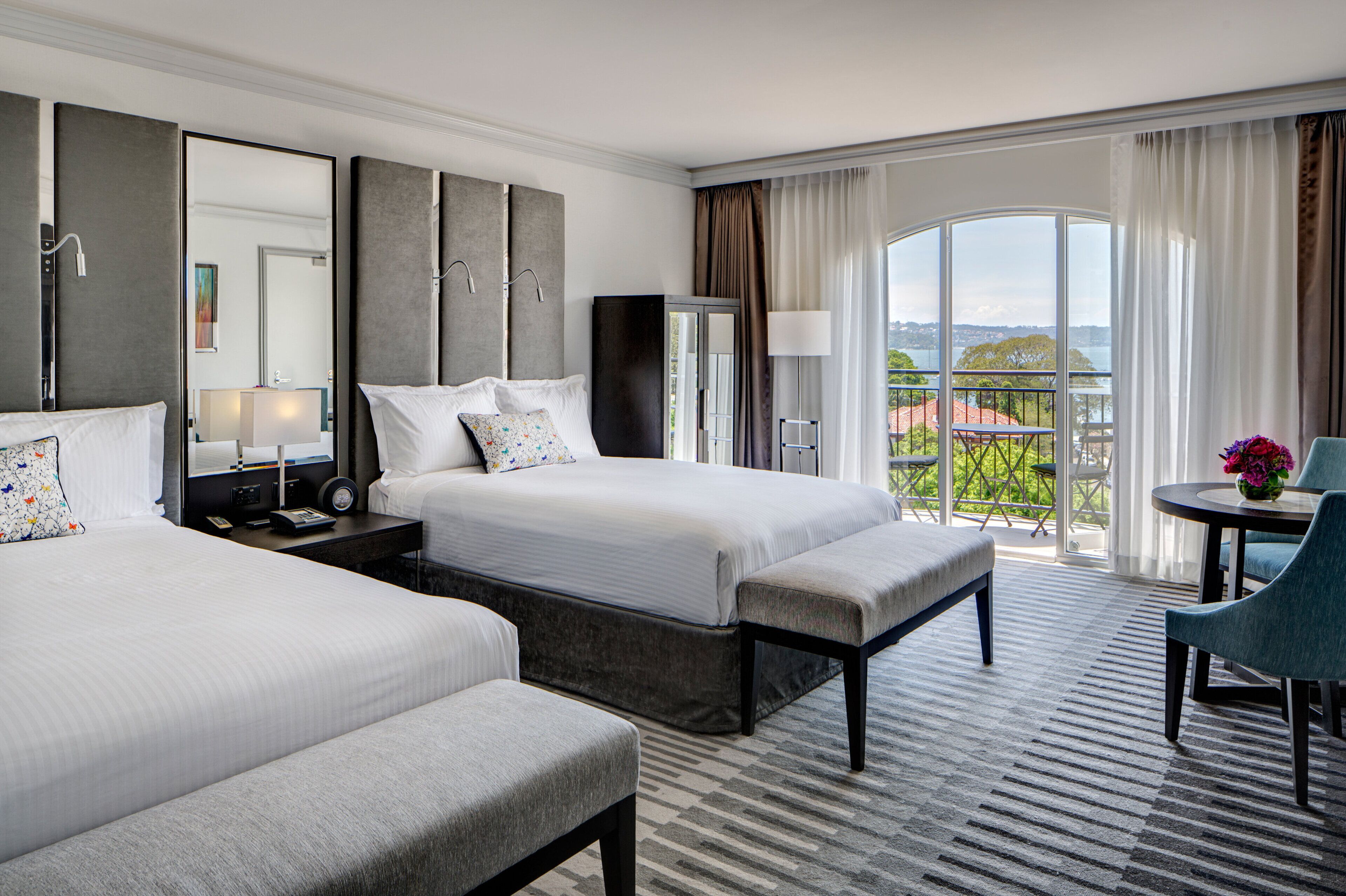 A hotel room with two beds, a desk, a chair, a balcony with a view, and a large mirror.