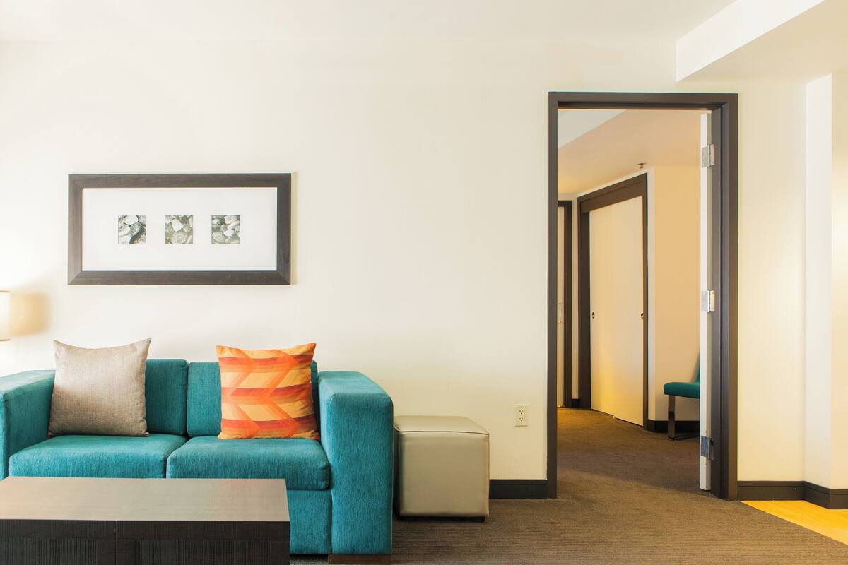 A teal sofa with two pillows, a small ottoman, and a framed picture on the wall.