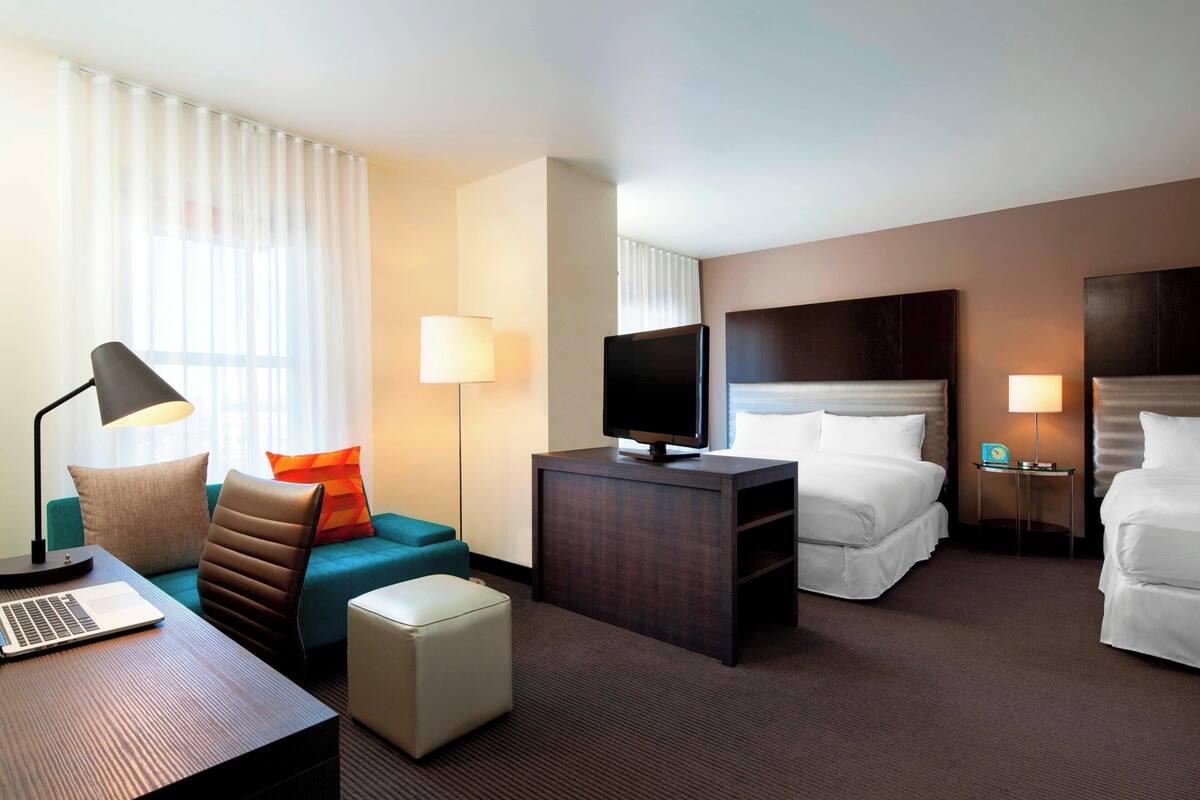 A hotel room with a bed, a desk with a laptop, a TV, a sofa, and a small table with a lamp.