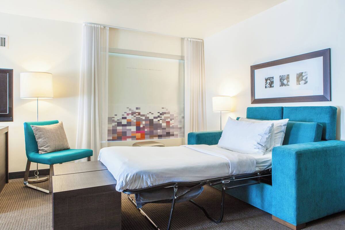 A hotel room with a bed, a blue armchair, a bedside table, a lamp, and a wall art piece.