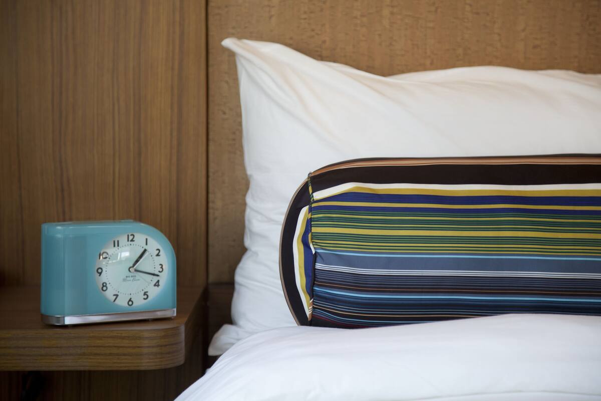 A bed with white sheets and a striped pillow, a blue clock on a wooden nightstand, and a wooden headboard.