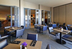 A hotel dining area with multiple tables and chairs, a kitchen in the background, and a view of the dining area through a glass partition.
