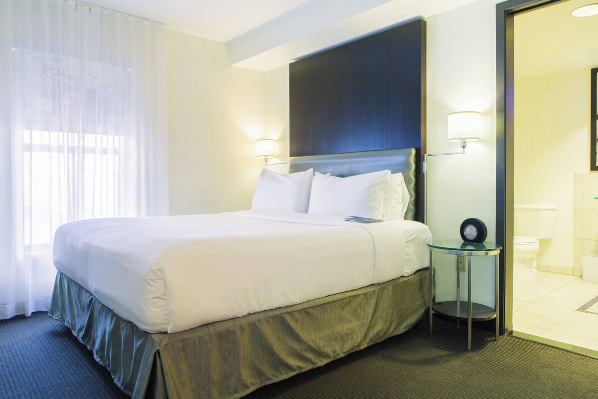 A hotel room with a large bed, a bedside table with a clock, and a bathroom visible through an open door.