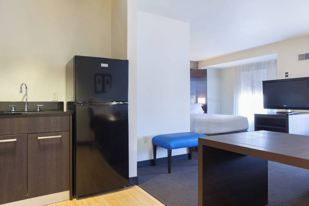 A compact hotel room with a kitchenette, a bed with a blue bench, a television, and a window with curtains.