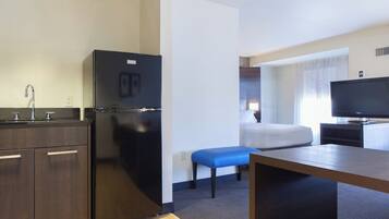 Quarto de hotel compacto com cozinha compacta, uma cama com um banco azul, televisão e janela com cortinas.