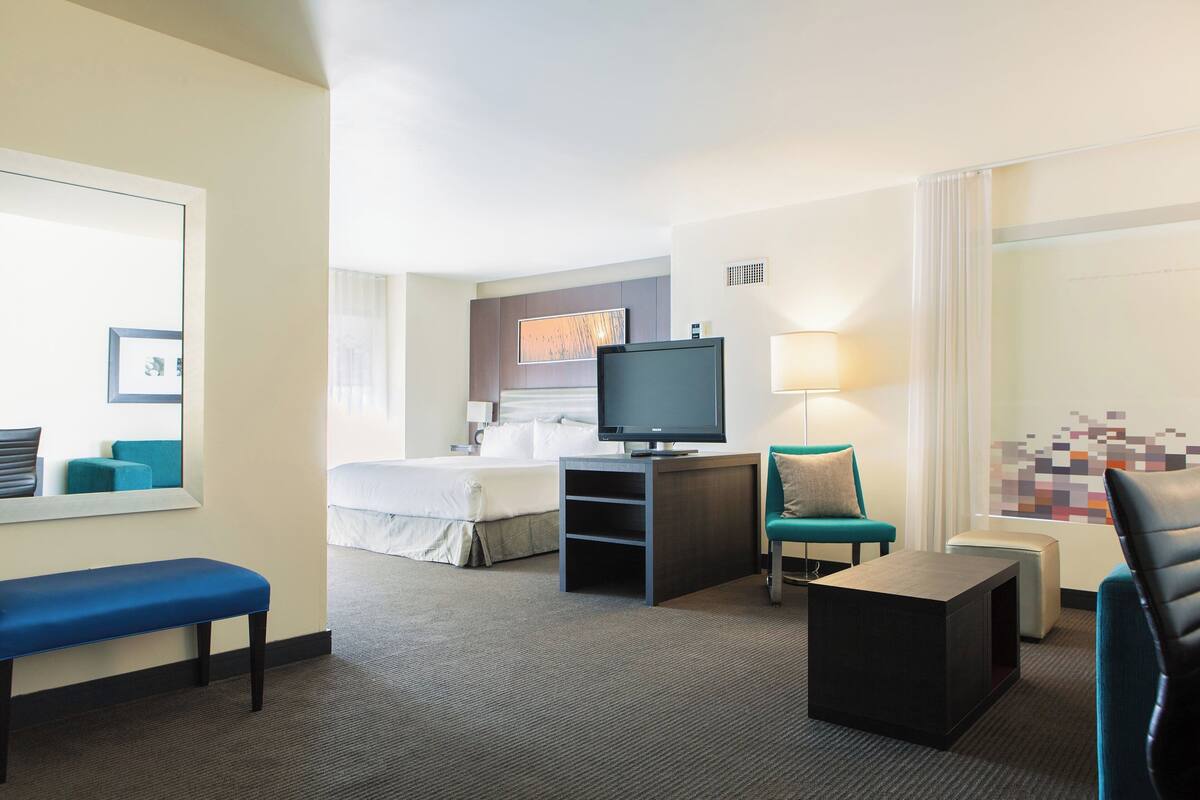 A hotel room with a bed, a desk with a flat-screen TV, a blue bench, a small table, and a chair.