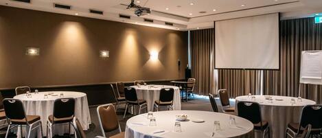 Un salón de conferencias con mesas redondas, sillas, un proyector y una pantalla.