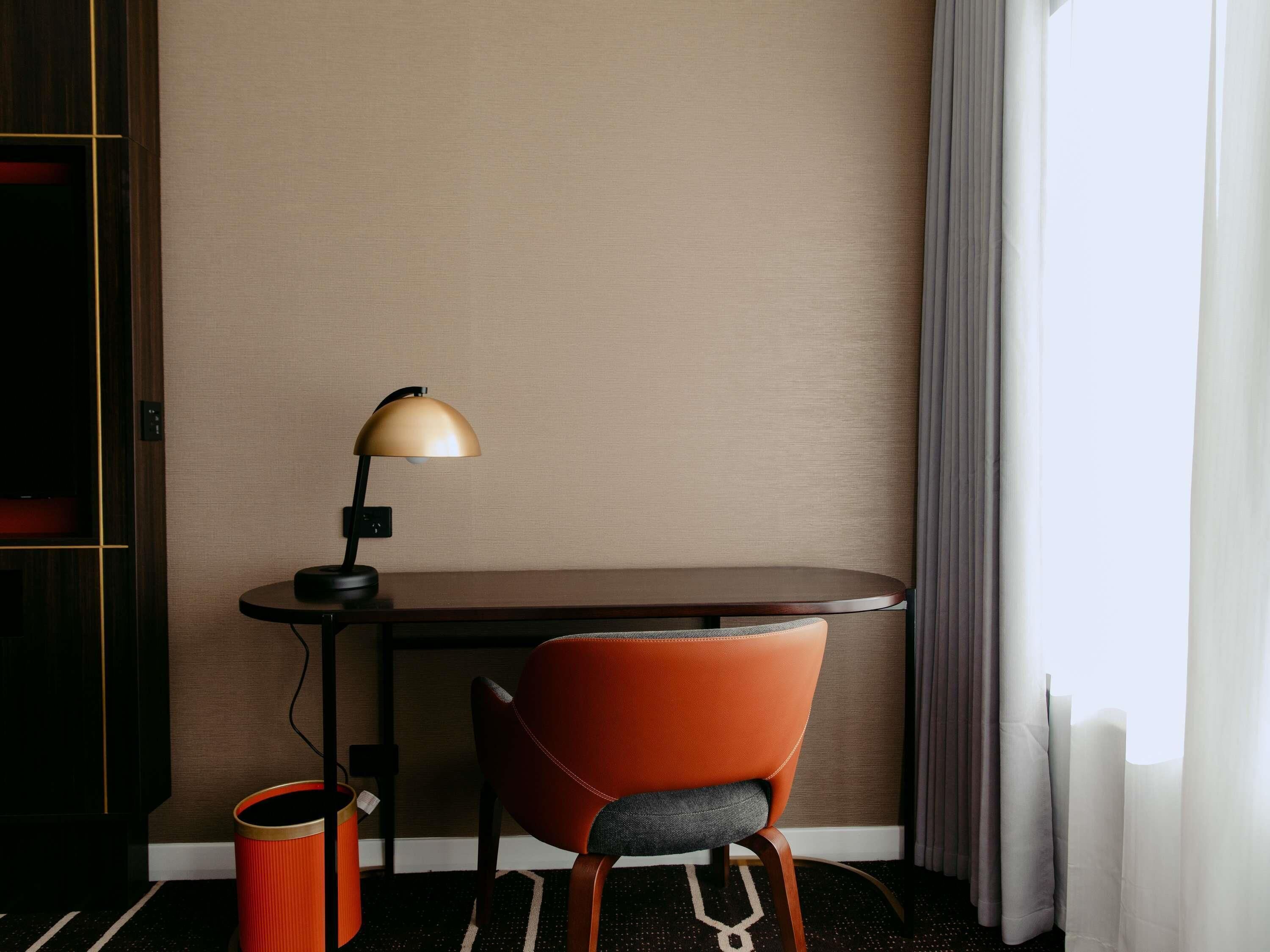 A hotel room with a desk, a chair, a lamp, a trash bin, and a window with curtains.