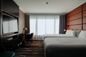 Habitación de hotel con dos camas, un escritorio con silla, televisor y un ventanal con cortinas.