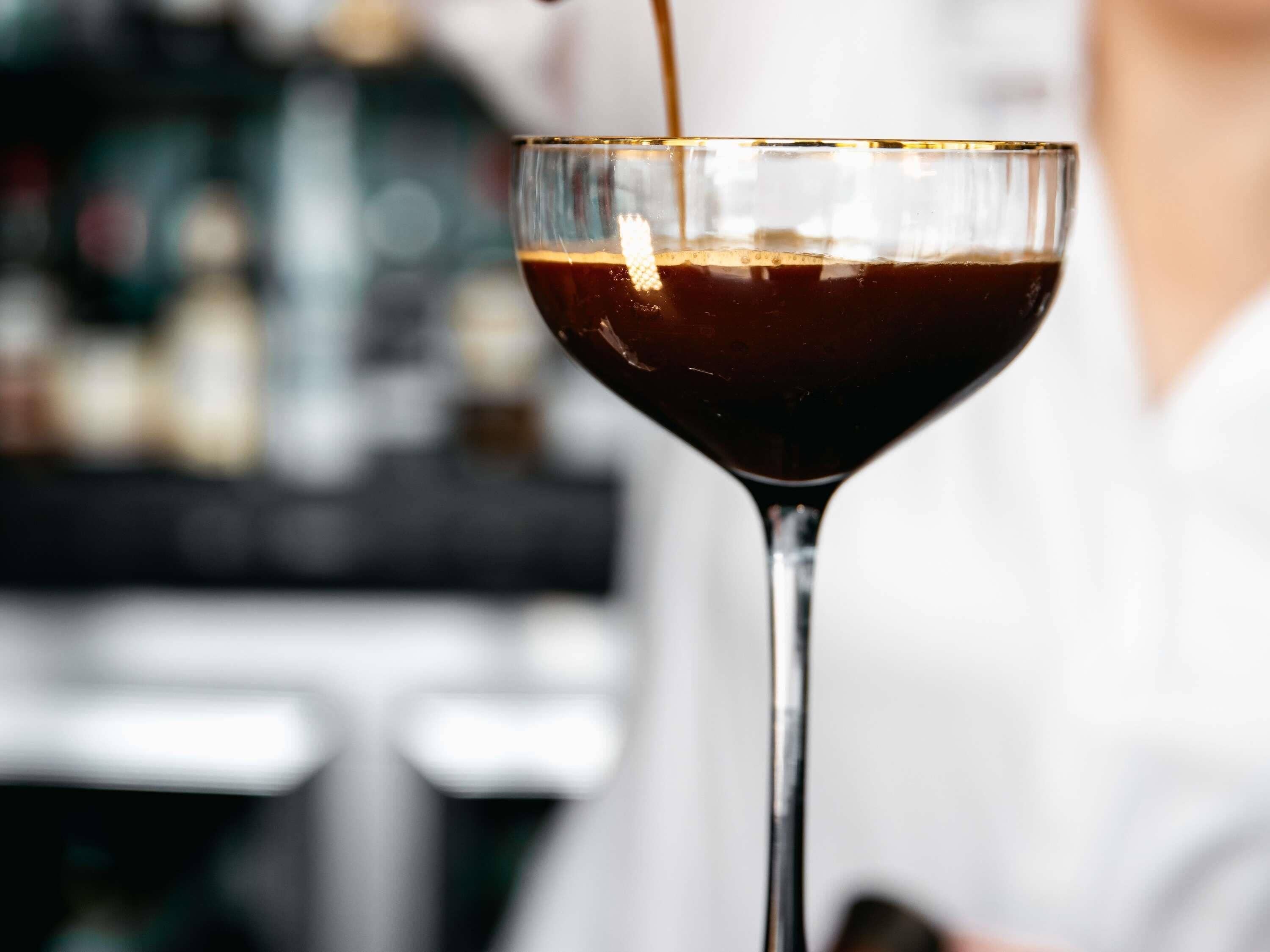 A martini glass filled with a dark liquid, likely a cocktail, being poured from a bottle.
